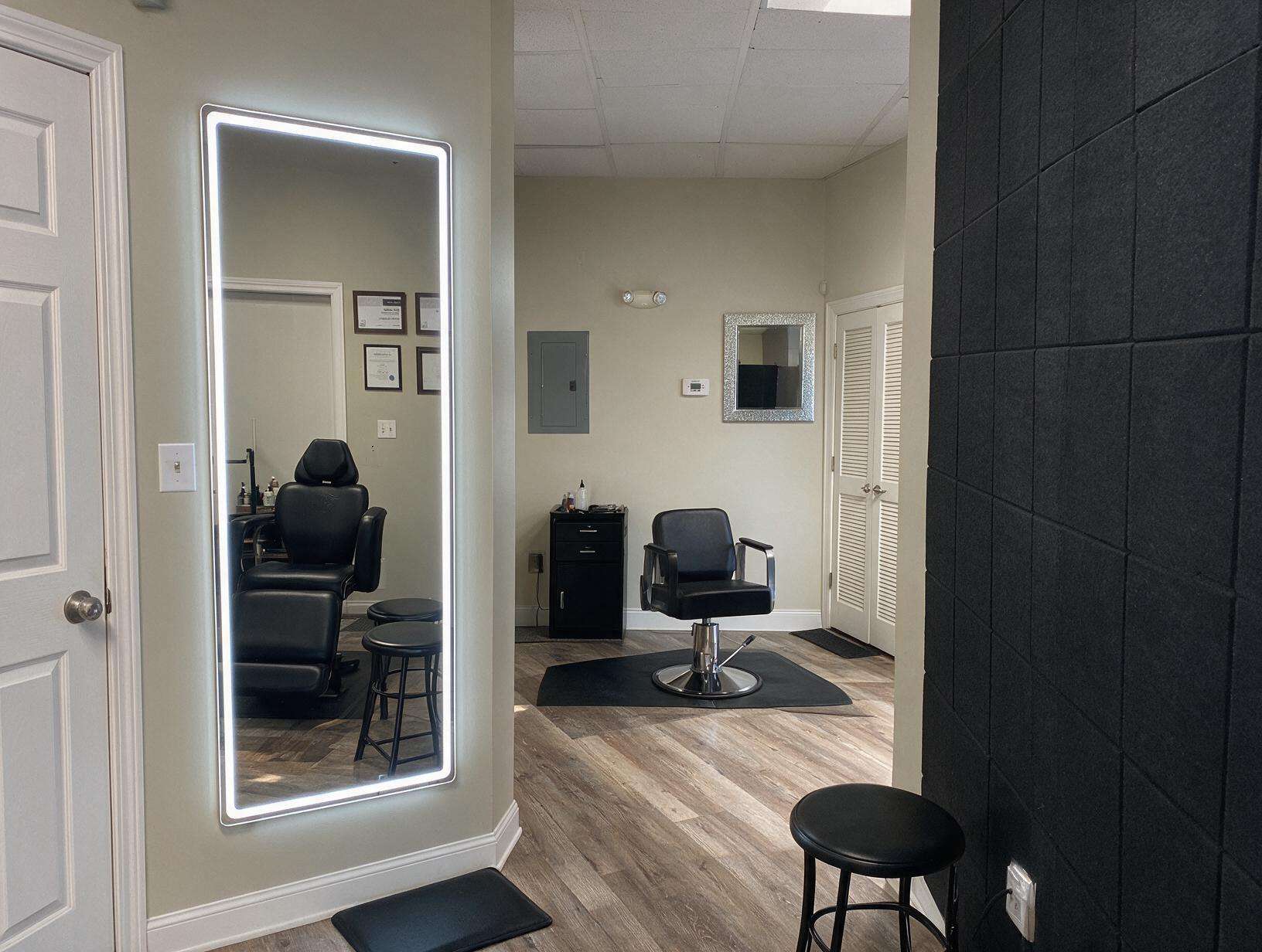 Empty studio room with a lit full-length mirror reflecting a black treatment chair and stools, a salon chair on a mat