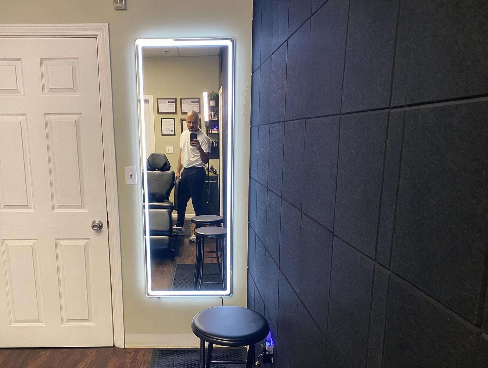 Man in a white T-shirt takes a phone selfie in an LED mirror, reflecting a black treatment chair