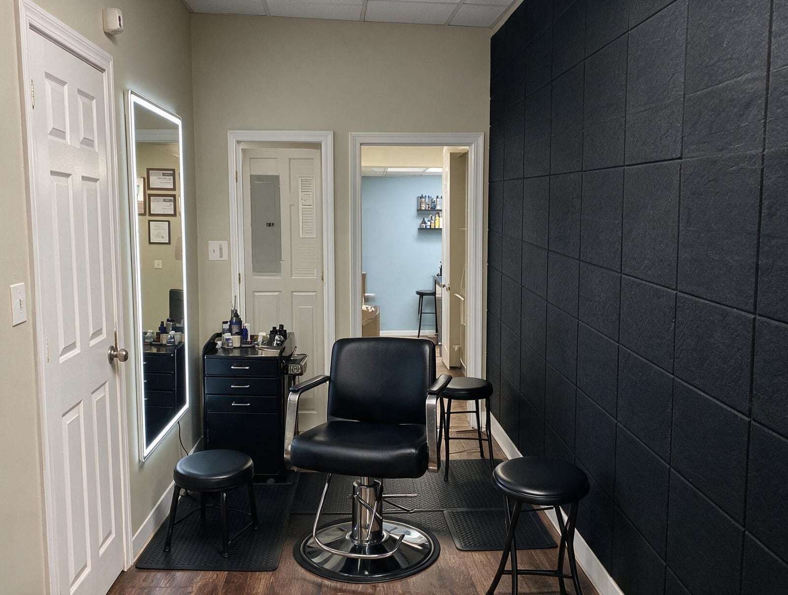 Empty treatment room with a black leather chair, black stools, a lit wall mirror, a black cabinet and a dark paneled wall.