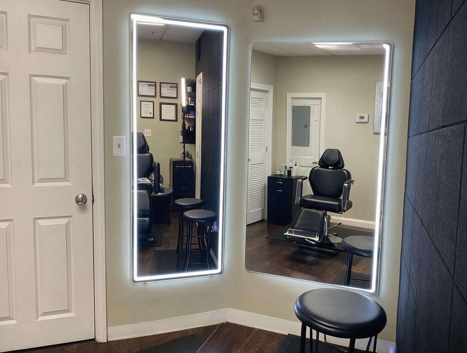 Empty treatment room with two lit wall mirrors, a black reclining chair, black stools, a cabinet and white doors