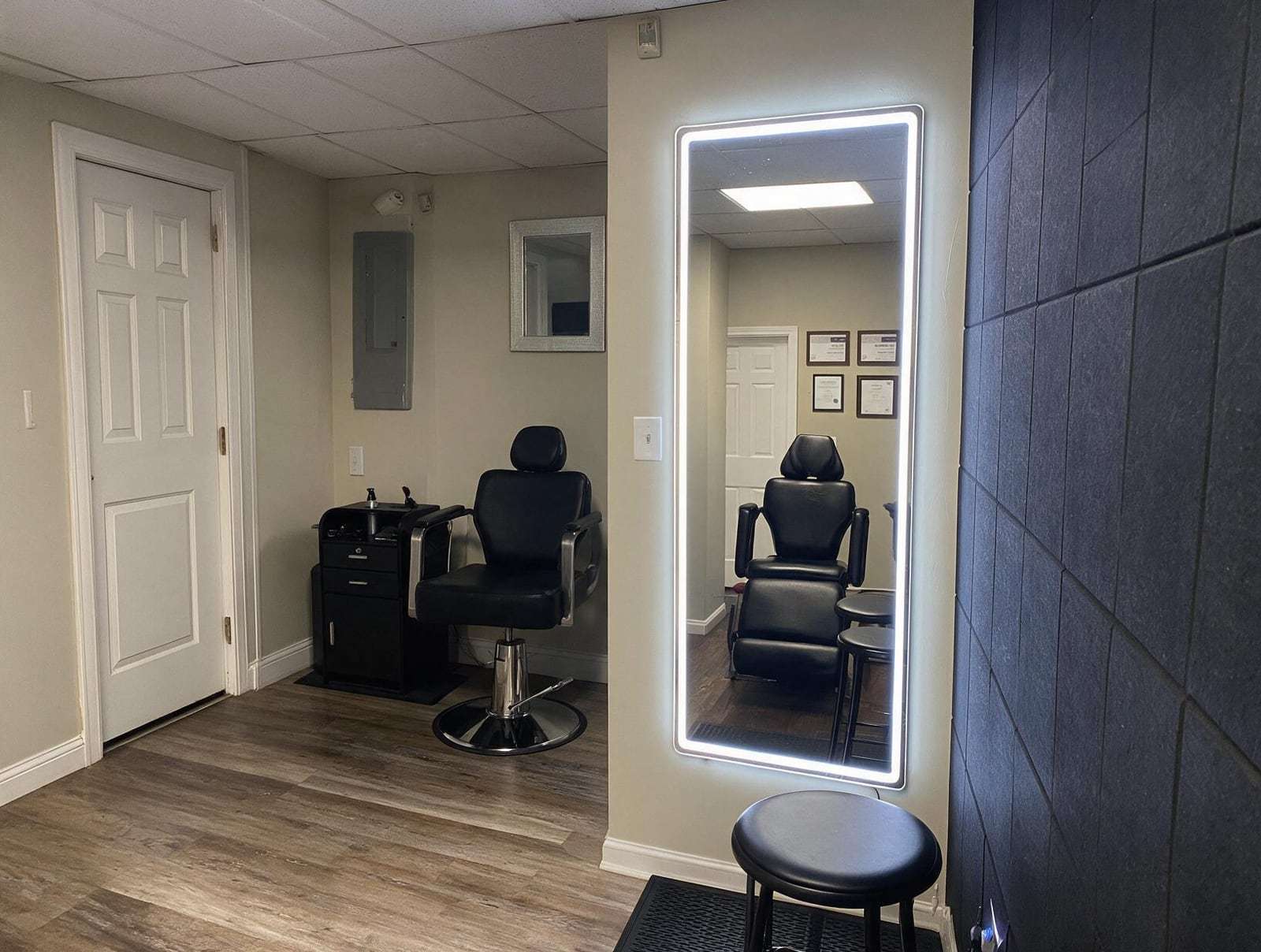 Empty studio room with a black salon chair and cabinet, a lit full-length mirror reflecting a treatment chair