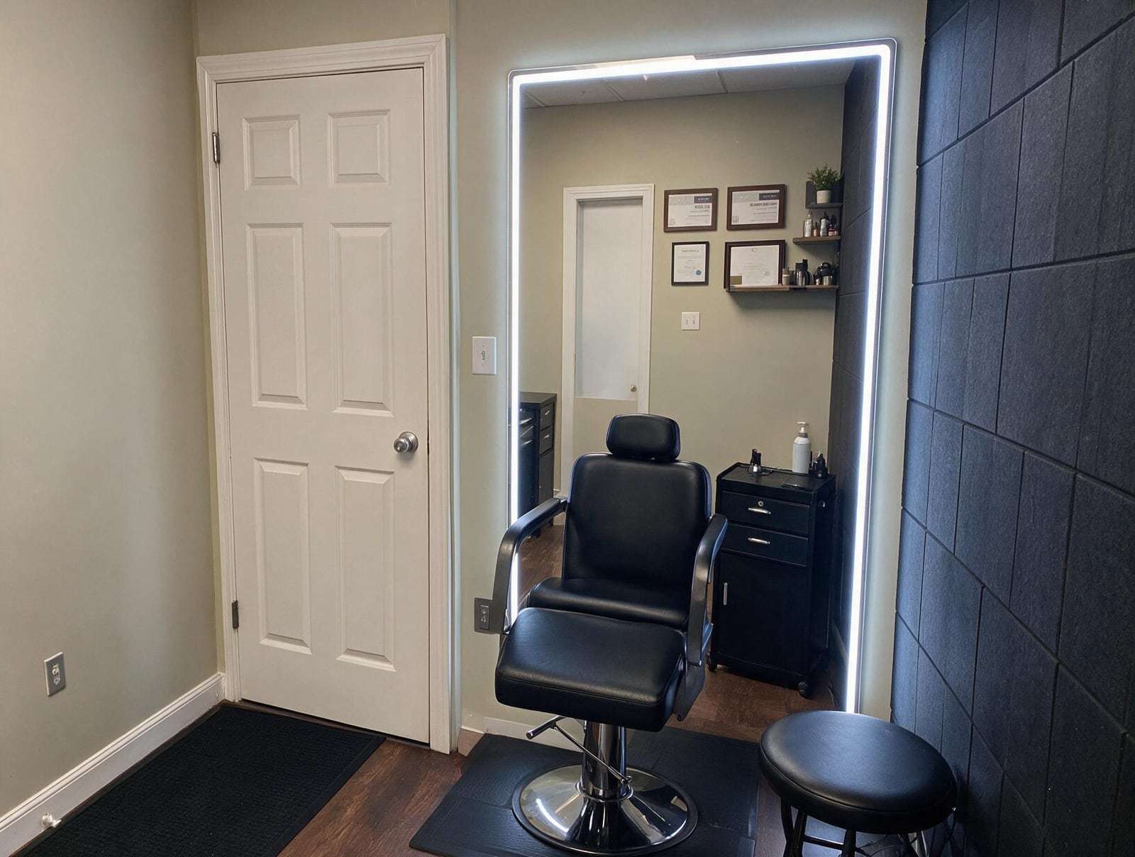 Empty treatment room with a black reclining chair, round stool, lit wall mirror