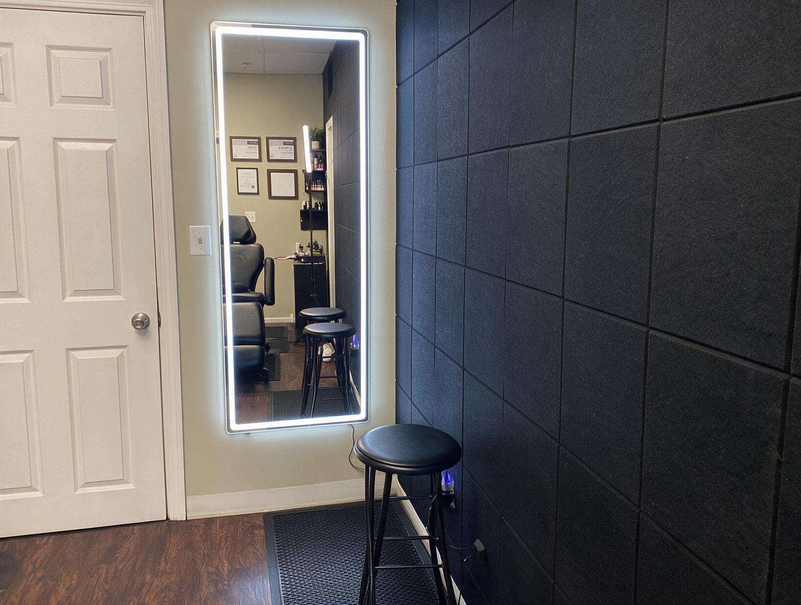 Empty treatment room corner with a lit full-length mirror reflecting a black treatment chair