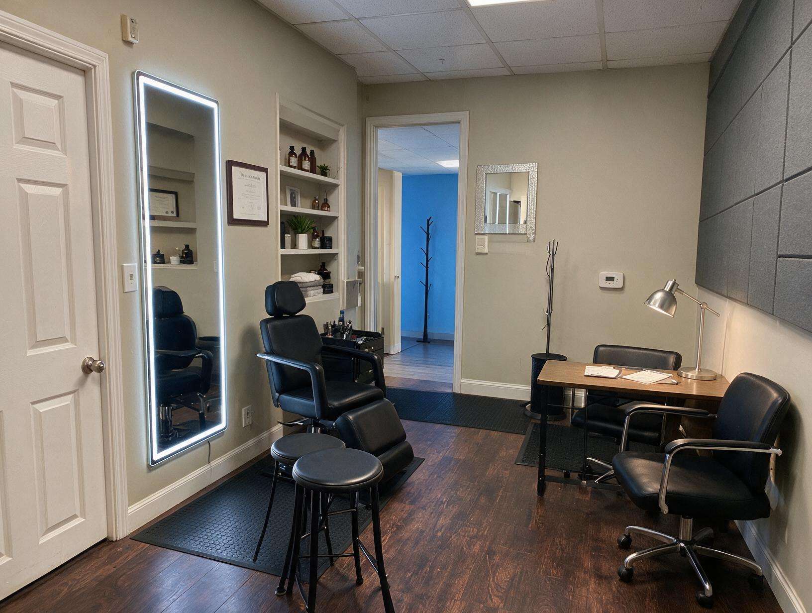 Empty treatment room with a black reclining chair, two stools, a lit wall mirror, built-in shelves