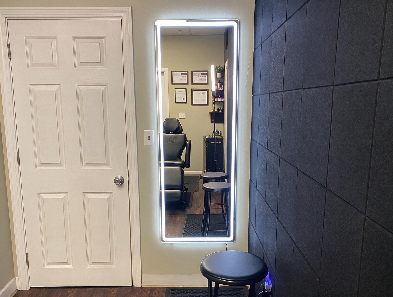 Empty treatment room with an LED-lit wall mirror reflecting a black reclining chair, stools and framed certificates
